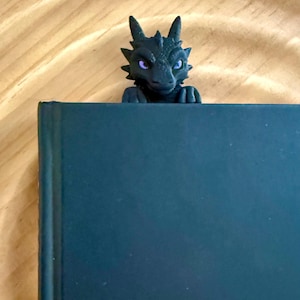 May include: A black dragon-shaped bookmark peeking over the top edge of a dark-colored book. The dragon figure features textured scales, pointed ears, and bright purple eyes, with its small front claws resting on the top edge of the book cover.