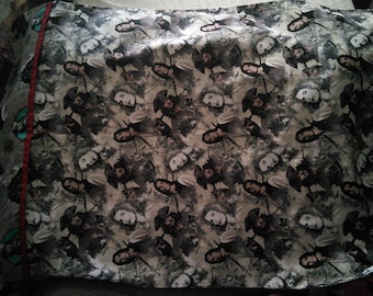 May include: A rectangular pillowcase with a repeating pattern of a woman's face and a figure in a hat. The design is in shades of gray, black, and white, with a red accent on the side. The pillowcase is likely made of fabric.