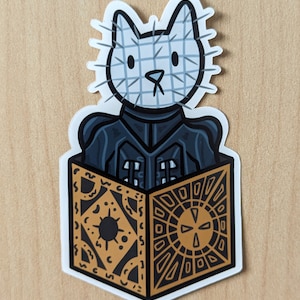 May include: A sticker of a white cat wearing a black and white mask with spikes, holding a golden box with a complex pattern. The box is open and the cat is looking out of it.