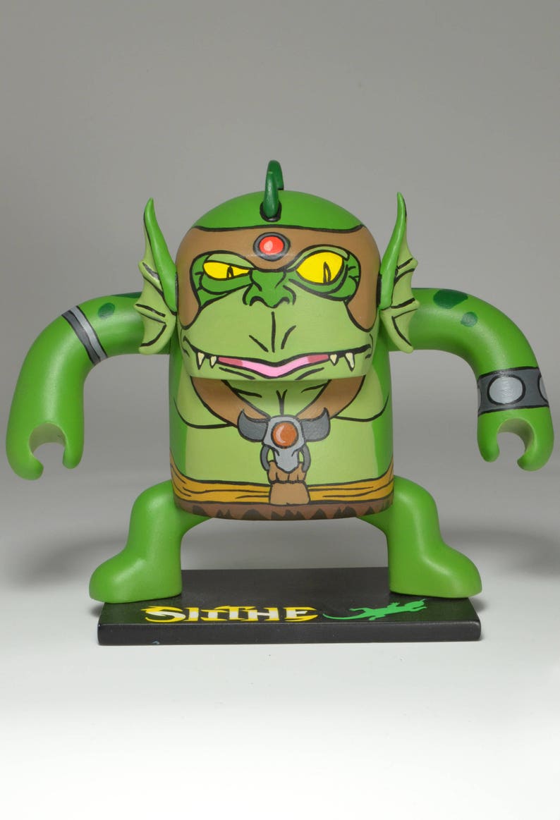 May include: Green and yellow hand-painted figurine of a mythical creature with a toothy grin, wearing a brown belt and a red jewel. The figurine is standing on a black base with the word "Slime" written in yellow.