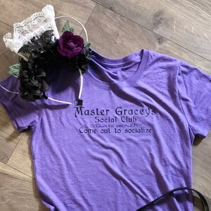 Puede incluir: Una camiseta morada con el texto "Master Gracey's Social Club Est. 1963 Come out to socialize" impreso en la parte delantera. Una bolsa de patr&oacute;n negro y gris con una correa negra est&aacute; en el suelo junto a la camiseta.