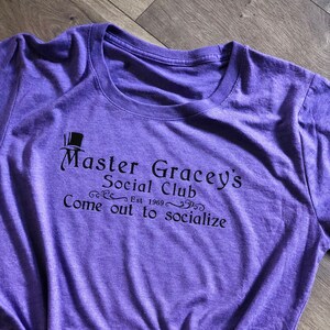 Puede incluir: Camiseta morada con un gr&aacute;fico negro que dice "Master Gracey's Social Club Est. 1969 Come out to socialize"