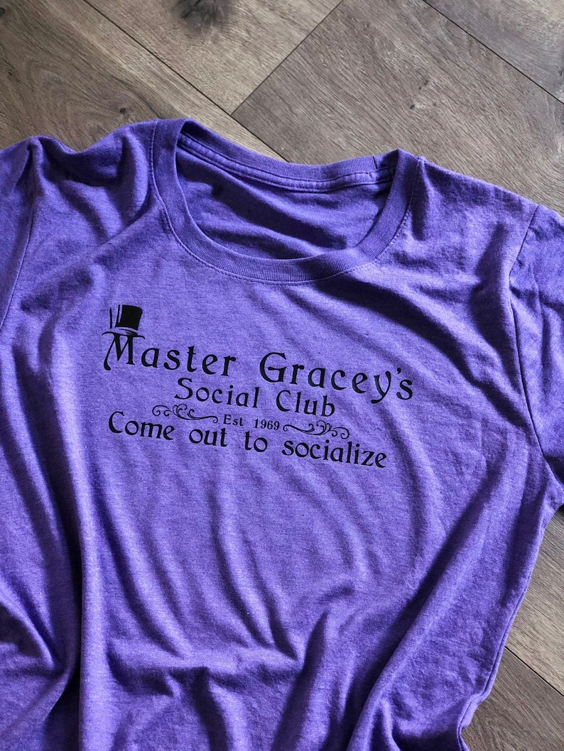 Puede incluir: Camiseta morada con un gr&aacute;fico negro que dice "Master Gracey's Social Club Est. 1969 Come out to socialize"