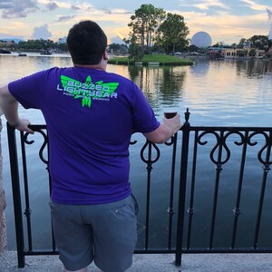 Puede incluir: Una persona con una camiseta morada con el texto "Buzzed Lightyear" y un logotipo verde. La persona est&aacute; de pie frente a una valla de metal negro y mirando hacia un cuerpo de agua. El cielo es azul y hay nubes en la distancia.