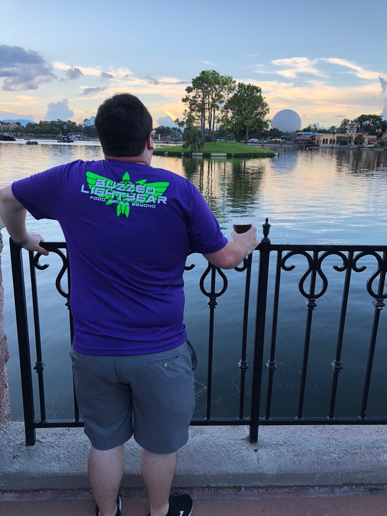 Puede incluir: Una persona con una camiseta morada con el texto "Buzzed Lightyear" y un logotipo verde. La persona est&aacute; de pie frente a una valla de metal negro y mirando hacia un cuerpo de agua. El cielo es azul y hay nubes en la distancia.