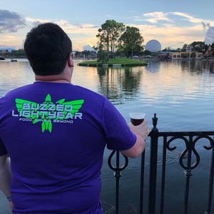 Puede incluir: Una persona con una camiseta morada con el texto "Buzzed Lightyear Food, Wine & Beyond" en letras verdes y blancas, de pie junto a una barandilla con vistas a un lago con una gran esfera blanca en el fondo.
