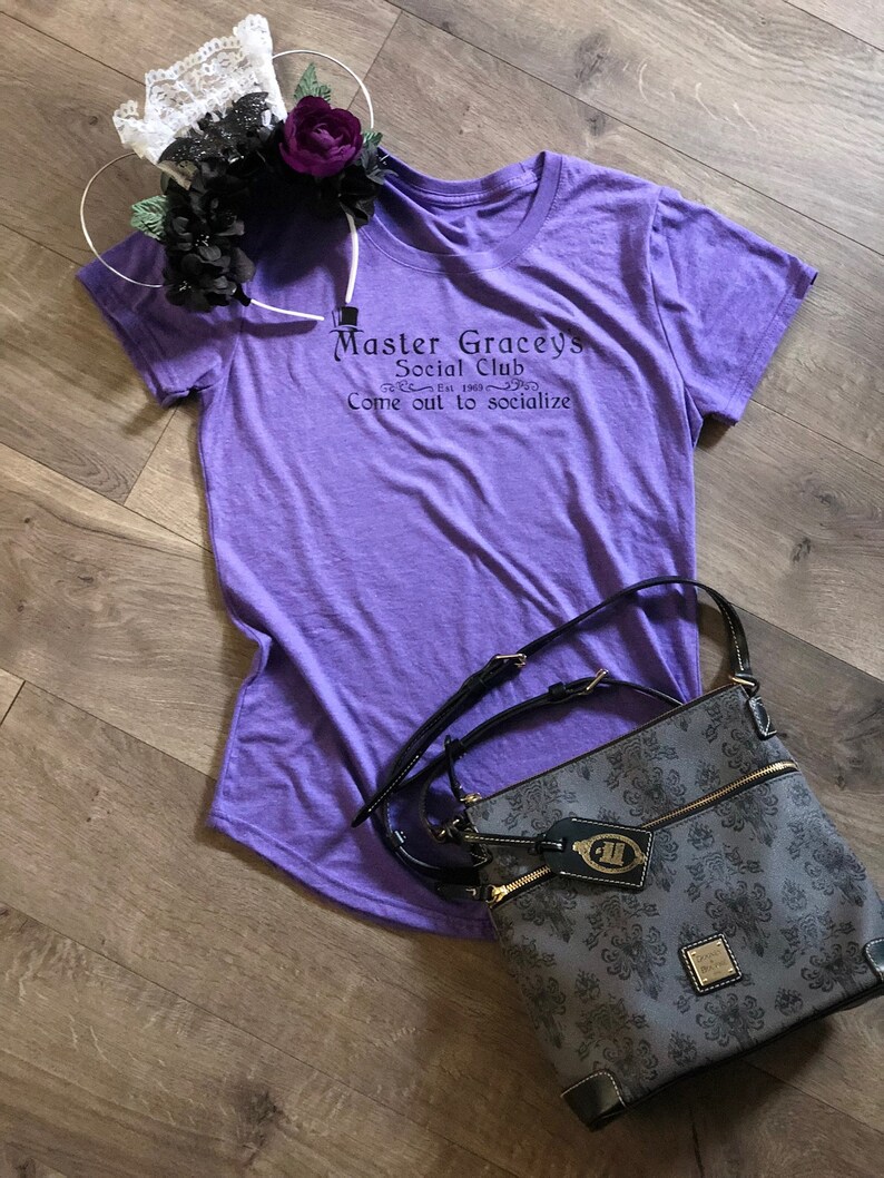 Puede incluir: Una camiseta morada con el texto "Master Gracey's Social Club Est. 1963 Come out to socialize" impreso en la parte delantera. Una bolsa de patr&oacute;n negro y gris con una correa negra est&aacute; en el suelo junto a la camiseta.