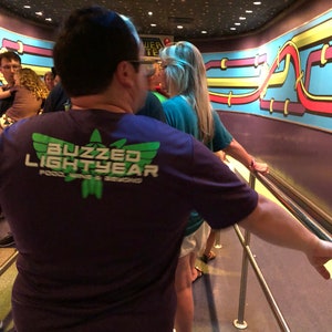 Puede incluir: Una persona lleva una camiseta morada con el texto "Buzzed Lightyear" y un logotipo verde. La persona est&aacute; de pie en una fila con otras personas. La pared detr&aacute;s de ellos tiene un dise&ntilde;o colorido con l&iacute;neas y curvas.