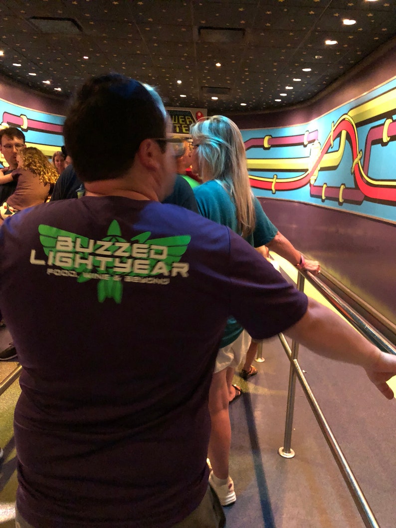 Puede incluir: Una persona lleva una camiseta morada con el texto "Buzzed Lightyear" y un logotipo verde. La persona est&aacute; de pie en una fila con otras personas. La pared detr&aacute;s de ellos tiene un dise&ntilde;o colorido con l&iacute;neas y curvas.