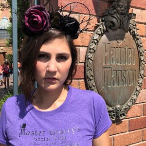 Puede incluir: Una mujer lleva una camiseta morada con el texto "Master Gracey's Social Club Come out to socialize" impreso en ella. Lleva una diadema floral negra y roja con ramas. La mujer est&aacute; de pie frente a una pared de ladrillo con un letrero que dice "The Haunted Mansion".