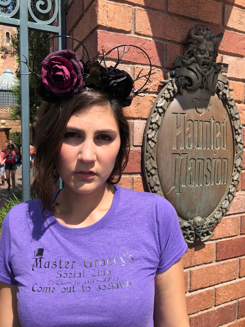 Puede incluir: Una mujer lleva una camiseta morada con el texto "Master Gracey's Social Club Come out to socialize" impreso en ella. Lleva una diadema floral negra y roja con ramas. La mujer est&aacute; de pie frente a una pared de ladrillo con un letrero que dice "The Haunted Mansion".