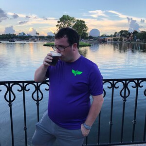 Puede incluir: Una persona con una camisa morada con un logotipo verde y pantalones cortos grises est&aacute; de pie junto a una barandilla y bebiendo de una taza negra. La persona est&aacute; de pie frente a un cuerpo de agua con una gran esfera en el fondo.