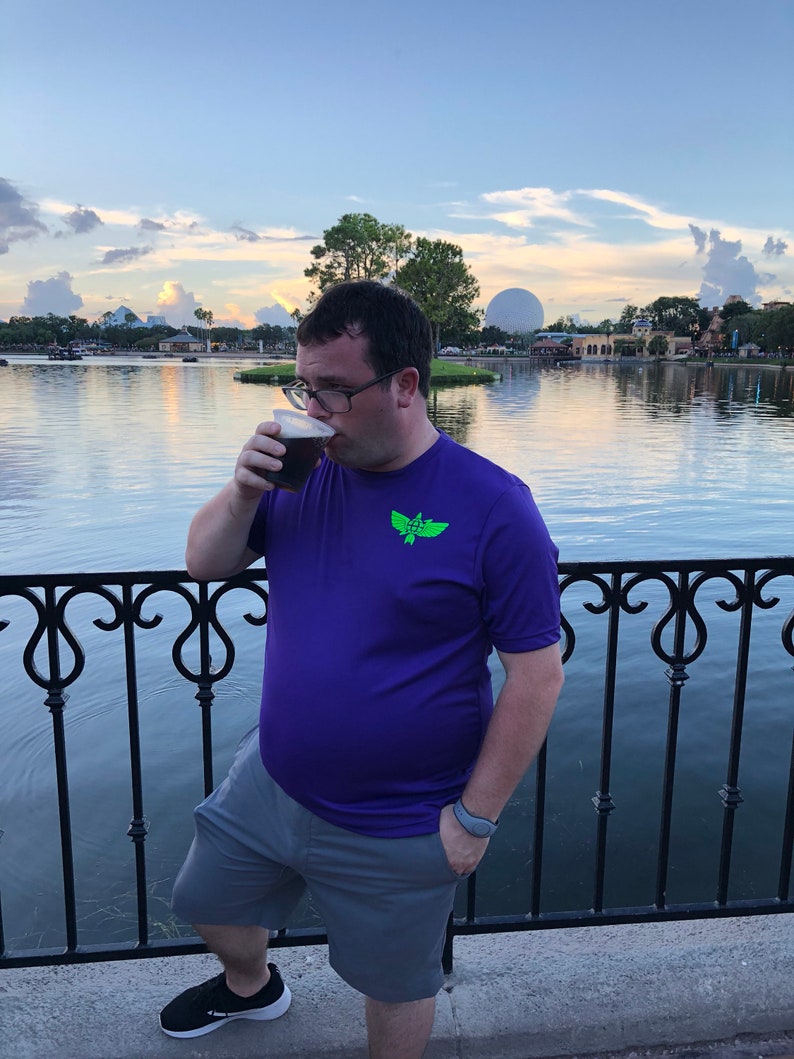 Puede incluir: Una persona con una camisa morada con un logotipo verde y pantalones cortos grises est&aacute; de pie junto a una barandilla y bebiendo de una taza negra. La persona est&aacute; de pie frente a un cuerpo de agua con una gran esfera en el fondo.