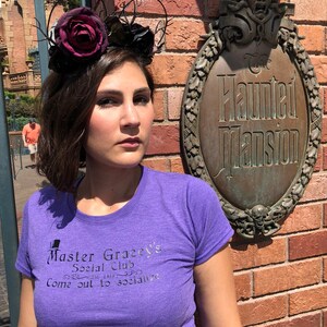 Puede incluir: Una mujer con una camiseta morada con el texto "Master Gracey's Social Club Come out to socialize" impreso en ella. Est&aacute; de pie frente a una pared de ladrillo con un letrero que dice "The Haunted Mansion". Lleva una diadema floral negra y burdeos.