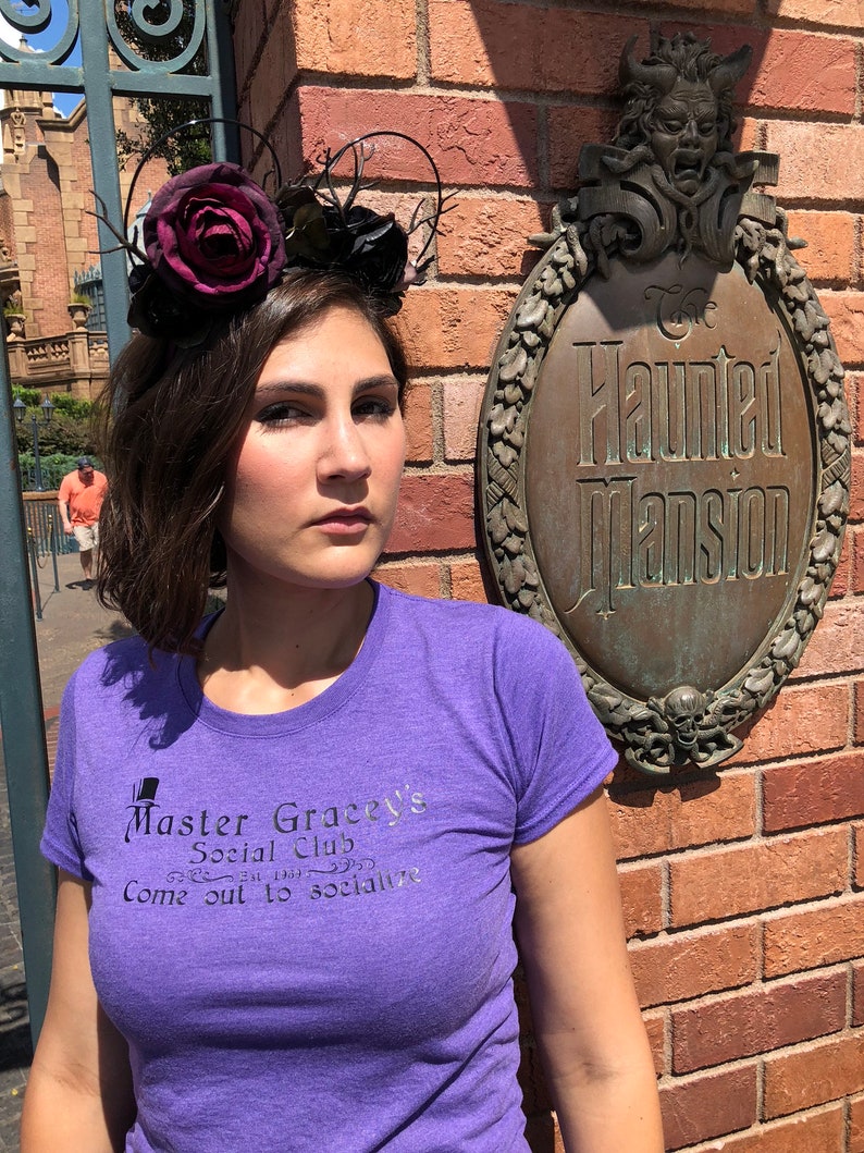 Puede incluir: Una mujer con una camiseta morada con el texto "Master Gracey's Social Club Come out to socialize" impreso en ella. Est&aacute; de pie frente a una pared de ladrillo con un letrero que dice "The Haunted Mansion". Lleva una diadema floral negra y burdeos.