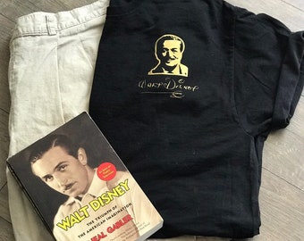May include: A black t-shirt with a gold silhouette of Walt Disney and his signature. A beige pair of shorts and a book titled "Walt Disney: The Triumph of the American Imagination" by Neal Gabler are also pictured.