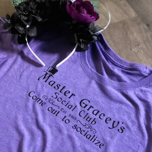Puede incluir: Camiseta morada con texto negro que dice "Master Gracey's Social Club Est. 1969 Come out to socialize".