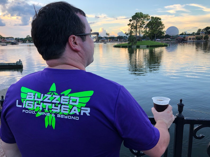 Puede incluir: Una persona con una camiseta morada con el texto "Buzzed Lightyear Food, Wine & Beyond" en verde y blanco. La persona sostiene una taza de cerveza y mira hacia un lago con una gran esfera en el fondo.