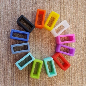 May include: A collection of colorful silicone watch band keepers in various colors, including black, orange, yellow, white, pink, purple, red, green, and blue. Each keeper has a rectangular shape with a rectangular opening and a glittery interior.