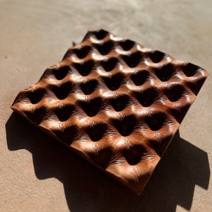 Könnte beinhalten: Eine braune, strukturierte, quadratische Fliese mit einem sich wiederholenden Muster aus erhabenen Kreisen. Die Fliese hat eine leicht gewellte Oberfläche und besteht aus einem Material, das wie Holz oder Leder aussieht.