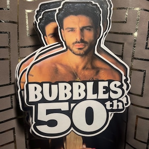 May include: A layered paper cutout cake topper featuring a photographic portrait of a man with dark hair and a beard, positioned above bold white text that reads BUBBLES 50th with a black outline, attached to a wooden stick for display.