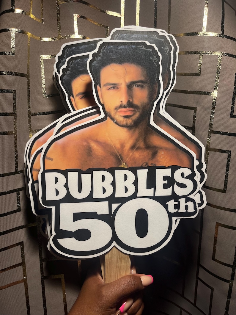 May include: A layered paper cutout cake topper featuring a photographic portrait of a man with dark hair and a beard, positioned above bold white text that reads BUBBLES 50th with a black outline, attached to a wooden stick for display.