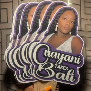 May include: A stack of die-cut paper fans featuring a photographic portrait of a person with long dark hair, wearing white earrings and a white top. The text "Crayani TAKES Bali" is printed in a bold purple and white font at the bottom of each fan.