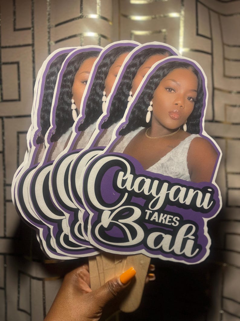 May include: A stack of die-cut paper fans featuring a photographic portrait of a person with long dark hair, wearing white earrings and a white top. The text "Crayani TAKES Bali" is printed in a bold purple and white font at the bottom of each fan.