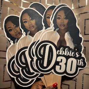 May include: A set of custom party cake toppers featuring a printed portrait of a person with long dark hair wearing a white shirt. The design includes the black and white text "Debbie's 30th" with a large stylized letter D. Each topper is attached to a wooden stick.