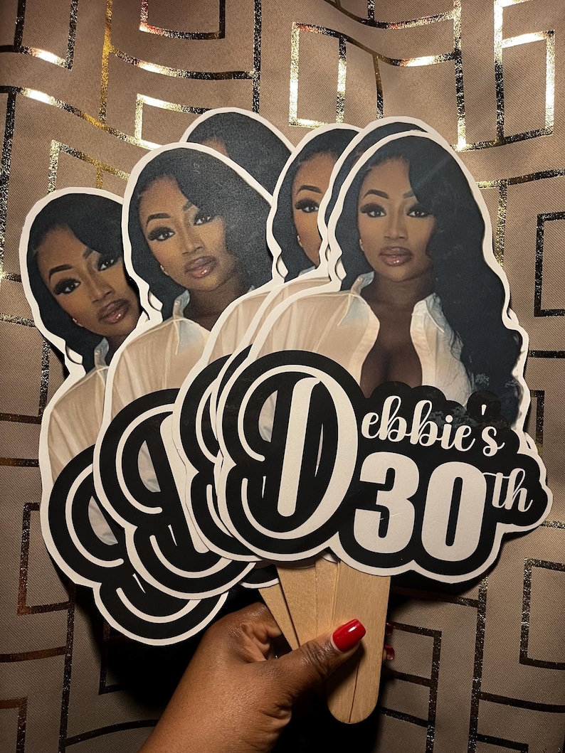 May include: A set of custom party cake toppers featuring a printed portrait of a person with long dark hair wearing a white shirt. The design includes the black and white text "Debbie's 30th" with a large stylized letter D. Each topper is attached to a wooden stick.
