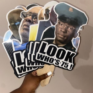 May include: A collection of handheld party props featuring die-cut photographic portraits of a person at different ages. Each cutout is attached to a wooden stick and displays the bold black and white text LOOK WHO'S 75! at the bottom edge.