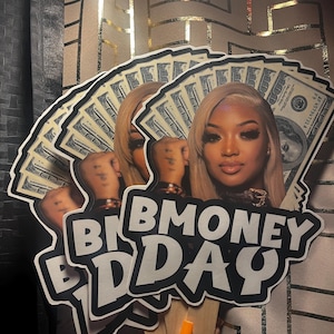 May include: A fan-shaped cutout featuring a portrait of a person with blonde hair, layered over a background of one hundred dollar bills. The text BMONEY DAY is printed in bold white letters with a black outline at the bottom of the cutout.