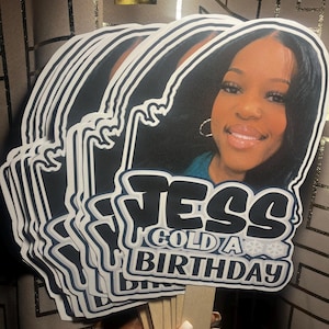 May include: A stack of custom handheld party fans featuring a photographic portrait of a person with dark hair and hoop earrings. The fans display the text JESS GOLD A BIRTHDAY in bold black and white lettering with snowflake accents on a stick.
