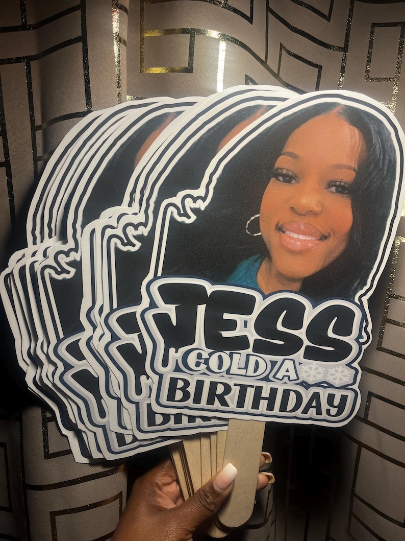 May include: A stack of custom handheld party fans featuring a photographic portrait of a person with dark hair and hoop earrings. The fans display the text JESS GOLD A BIRTHDAY in bold black and white lettering with snowflake accents on a stick.