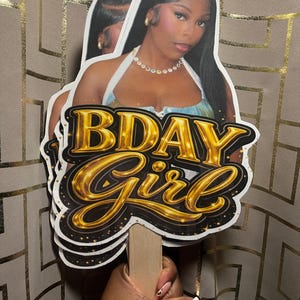 May include: A custom party prop featuring a photographic cutout of a person with long dark hair wearing a white halter top and a beaded necklace, layered over a black and gold graphic sign that reads BDAY Girl in bold, metallic lettering.