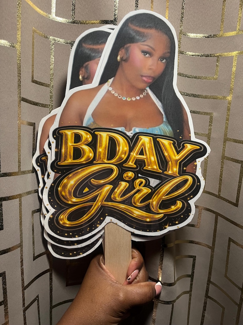 May include: A custom party prop featuring a photographic cutout of a person with long dark hair wearing a white halter top and a beaded necklace, layered over a black and gold graphic sign that reads BDAY Girl in bold, metallic lettering.