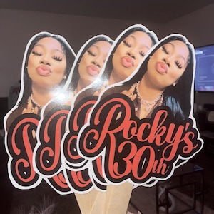 May include: A set of four identical handheld party props featuring a photographic portrait of a person with long dark hair and a pouting expression. Below the portrait, the text reads Rocky's 30th in a bold, red, cursive font with a black outline.