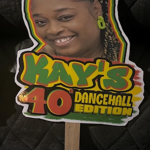 May include: A custom cake topper featuring a portrait of a person with braided hair and green earrings, surrounded by a red, yellow, and green border. The text reads KAY'S 40th DANCEHALL EDITION in red, yellow, and green lettering on a wooden stick.