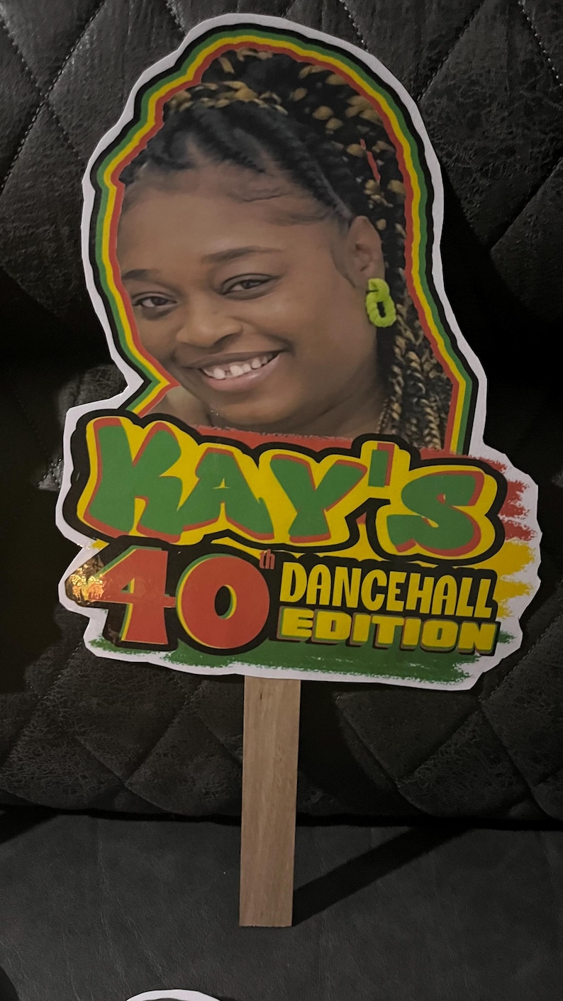 May include: A custom cake topper featuring a portrait of a person with braided hair and green earrings, surrounded by a red, yellow, and green border. The text reads KAY'S 40th DANCEHALL EDITION in red, yellow, and green lettering on a wooden stick.