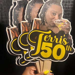 May include: A set of three handheld paper fans featuring a photographic print of a person holding a cocktail glass. The text "Terri's 50th" is printed in large yellow and black script and block lettering across the bottom of each fan.