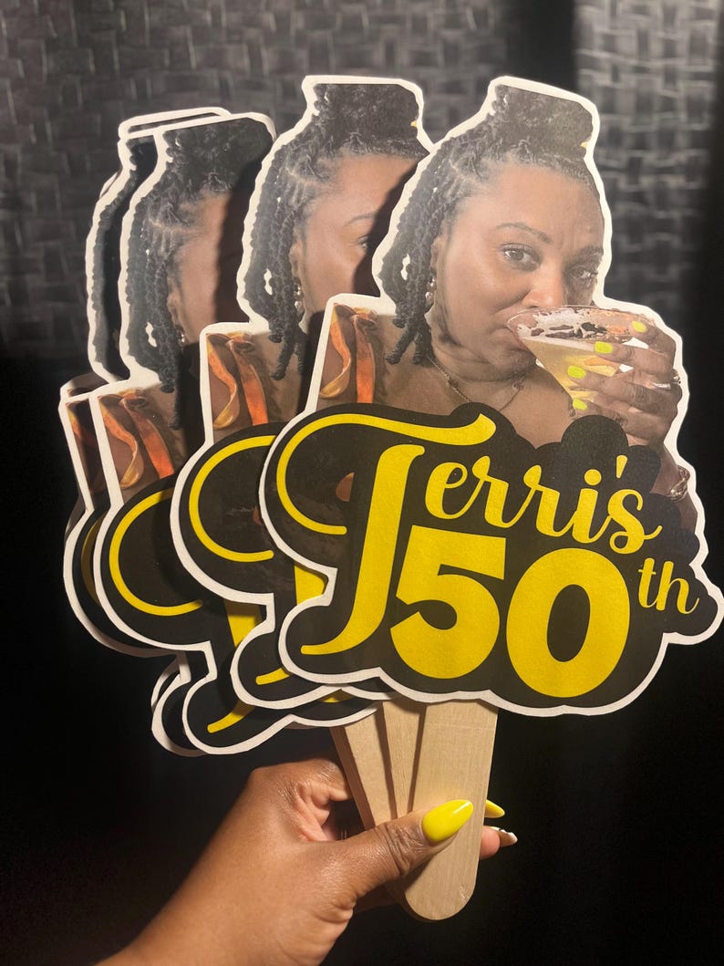 May include: A set of three handheld paper fans featuring a photographic print of a person holding a cocktail glass. The text "Terri's 50th" is printed in large yellow and black script and block lettering across the bottom of each fan.