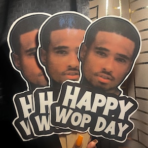 May include: Three identical handheld paper cutouts featuring a photographic portrait of a man with a mustache and goatee. Below the portrait, bold white text on a black background reads "HAPPY WOP DAY" in a sans-serif font. Each cutout has a wooden stick.
