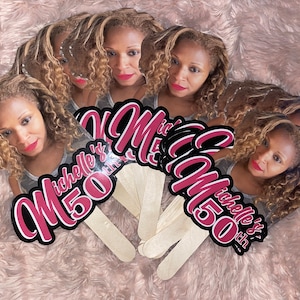 May include: A set of custom handheld fans featuring a photographic portrait of a person with curly blonde hair and red lipstick. The text Michelle's 50th is printed in a bold, dark pink font with a black outline on each fan, attached to wooden sticks.