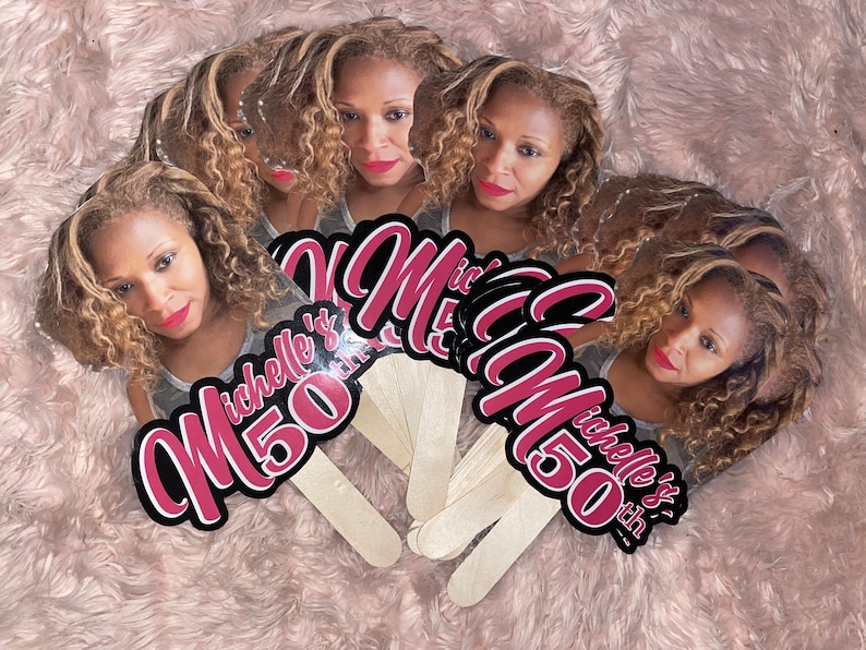 May include: A set of custom handheld fans featuring a photographic portrait of a person with curly blonde hair and red lipstick. The text Michelle's 50th is printed in a bold, dark pink font with a black outline on each fan, attached to wooden sticks.