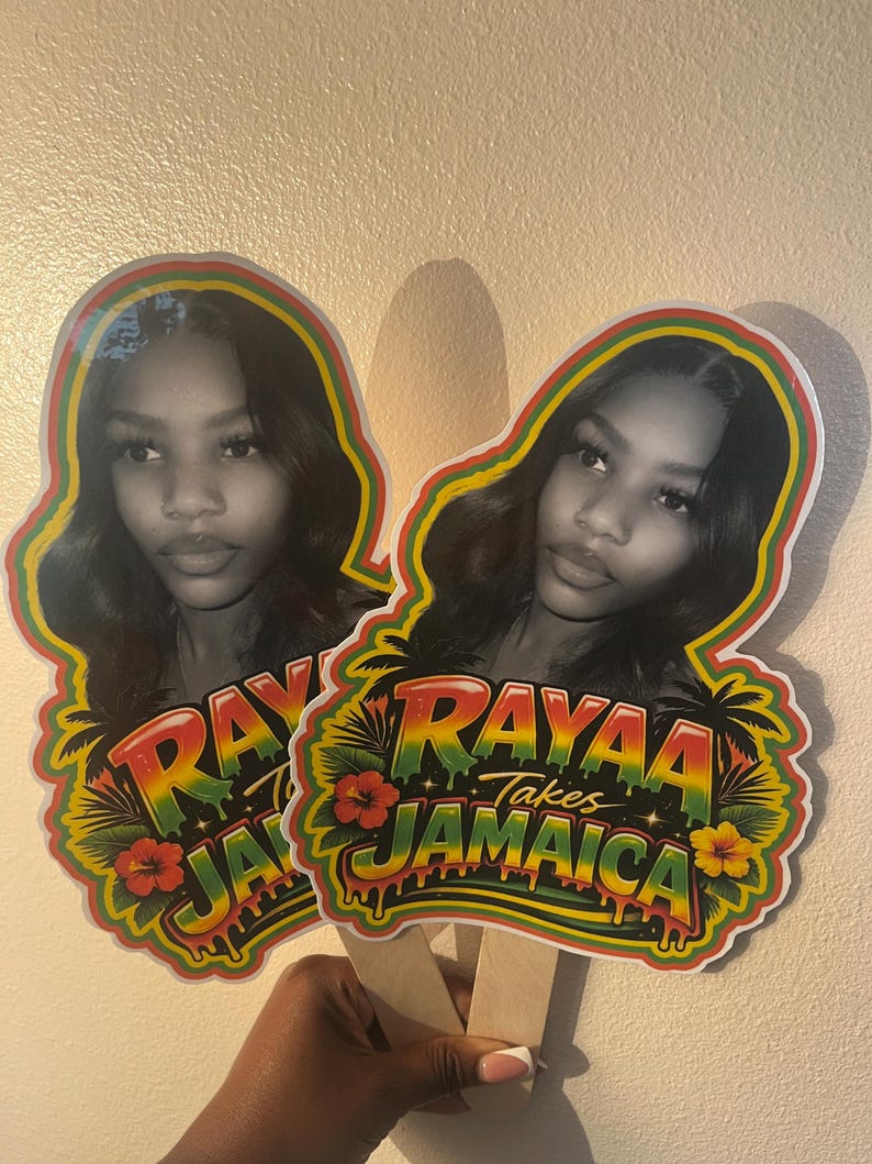 May include: Two custom handheld fans featuring a black and white portrait of a person. The design includes the text "RAYAA Takes JAMAICA" in a red, yellow, and green gradient font, surrounded by tropical palm tree and hibiscus flower graphics.