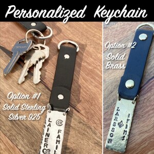 May include: Personalized keychain with two options: solid sterling silver 925 or solid brass. The keychain features a black leather strap and a rectangular tag with the text "FAMILY" and "LAINER" engraved on it. The second option features a rectangular tag with the text "LARSSON" and "BMS" engraved on it.