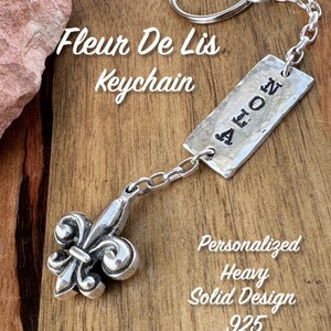 May include: A silver Fleur De Lis keychain with a rectangular tag that reads "NOLA". The keychain has a solid design and is marked "925". The text "Fleur De Lis Keychain" and "Personalized Heavy Solid Design" are also visible.