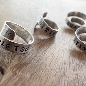 May include: Four silver rings with different words engraved on them. The rings are on a wooden surface. The words on the rings are "This Too", "Positive", "Be", and "R".