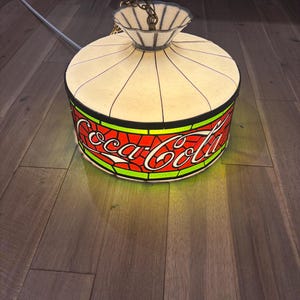 Puede incluir: Una lámpara colgante vintage de Coca-Cola con una pantalla blanca y crema. La lámpara presenta el logotipo de Coca-Cola en rojo con un borde verde y amarillo. La lámpara está suspendida por una cadena.