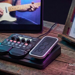 Op de afbeelding: Een zwarte Zoom G2X Four gitaar multi-effect processor met een aangesloten pedaal, een tablet met een gitarist en bladmuziek op een houten standaard. De opstelling staat op een houten oppervlak.
